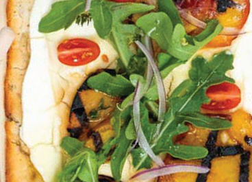 A table of food featuring cheese and vibrant vegetables on flat bread, pizza crust, and rolls.